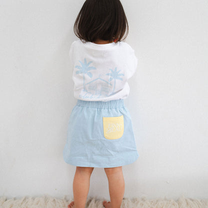 Child wearing a white shirt with blue floral patterns and a light blue skirt with a yellow pocket against a plain background.