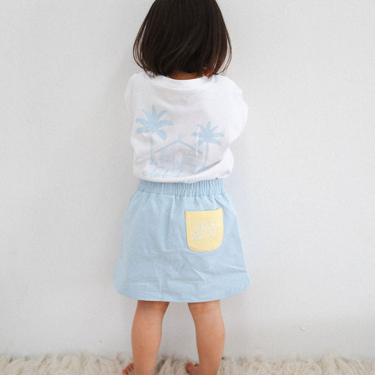 Child wearing a white shirt with blue floral patterns and a light blue skirt with a yellow pocket against a plain background.