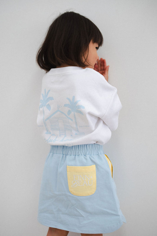 Child wearing a white hoodie with snowflake designs and a light blue skirt with a yellow pocket on a plain background