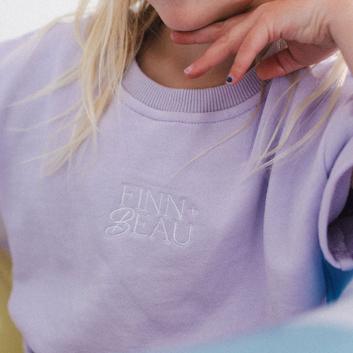 Child wearing a light purple sweatshirt with 'FINN & ELLA' branding, sitting on a light blue surface.