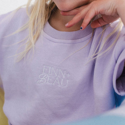 Child wearing a light purple sweatshirt with 'FINN & ELLA' branding, sitting on a light blue surface.