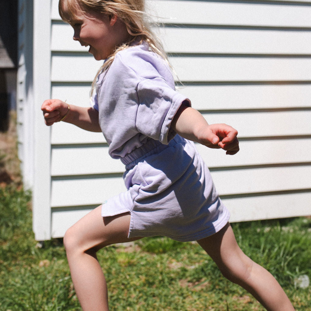 Child in a light purple outfit running outdoors with a white building in the background