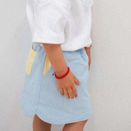 Child wearing a white shirt and light blue skirt with a red bracelet on a plain background